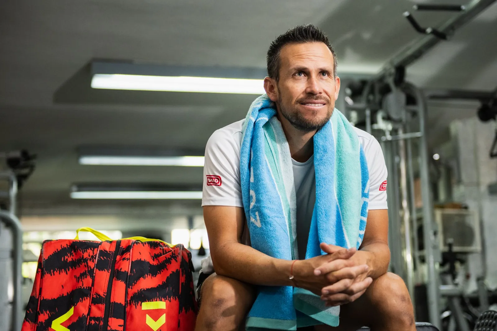 Miguel rests in the gym with a towel around his neck, next to his racket bag.