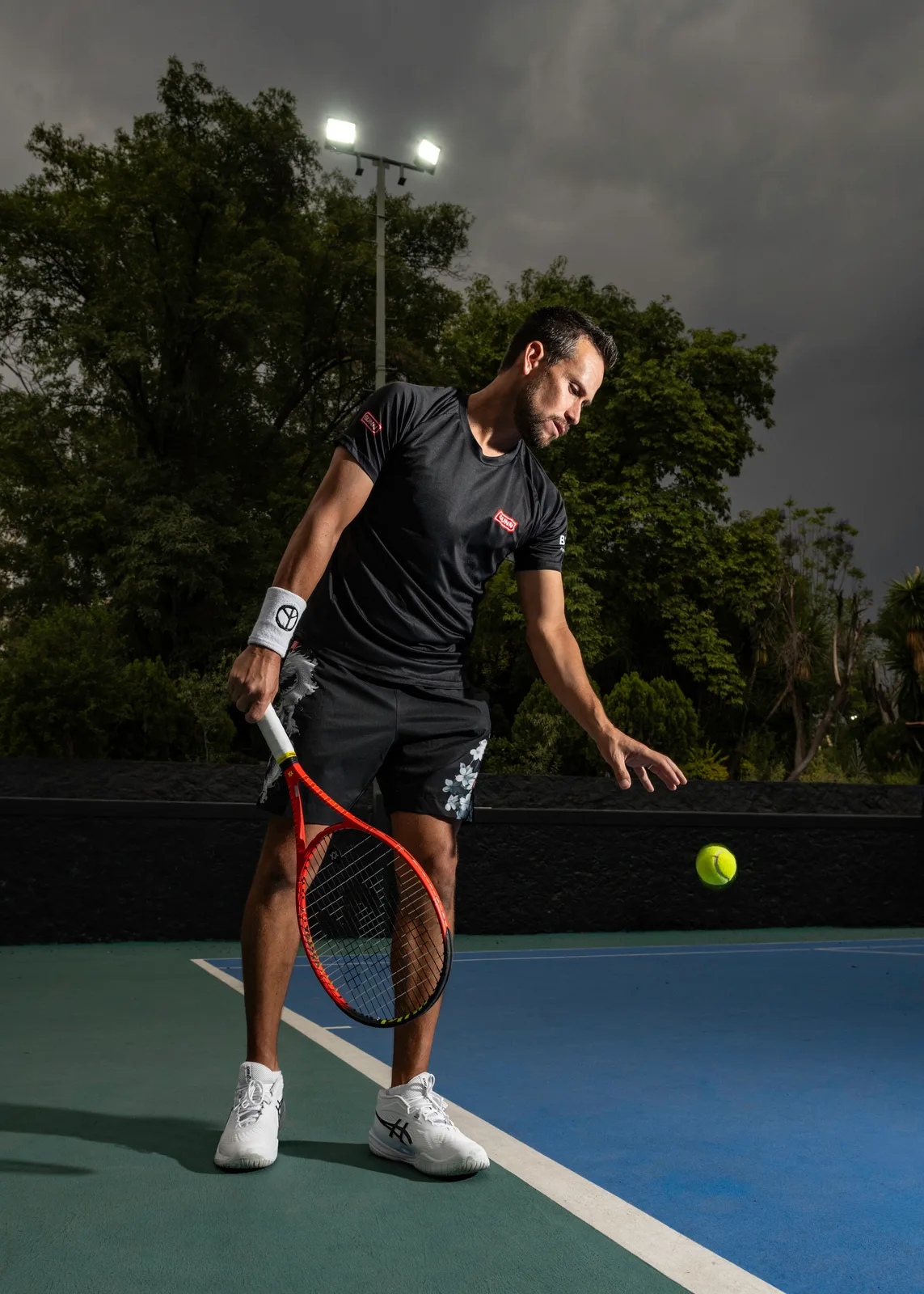 Miguel bounces the ball before serving, at night under the court lights.