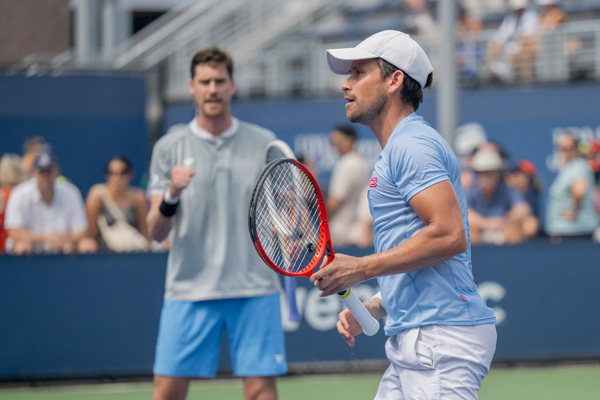 Miguel, in a white cap and holding his racket, on court beside his doubles partner.