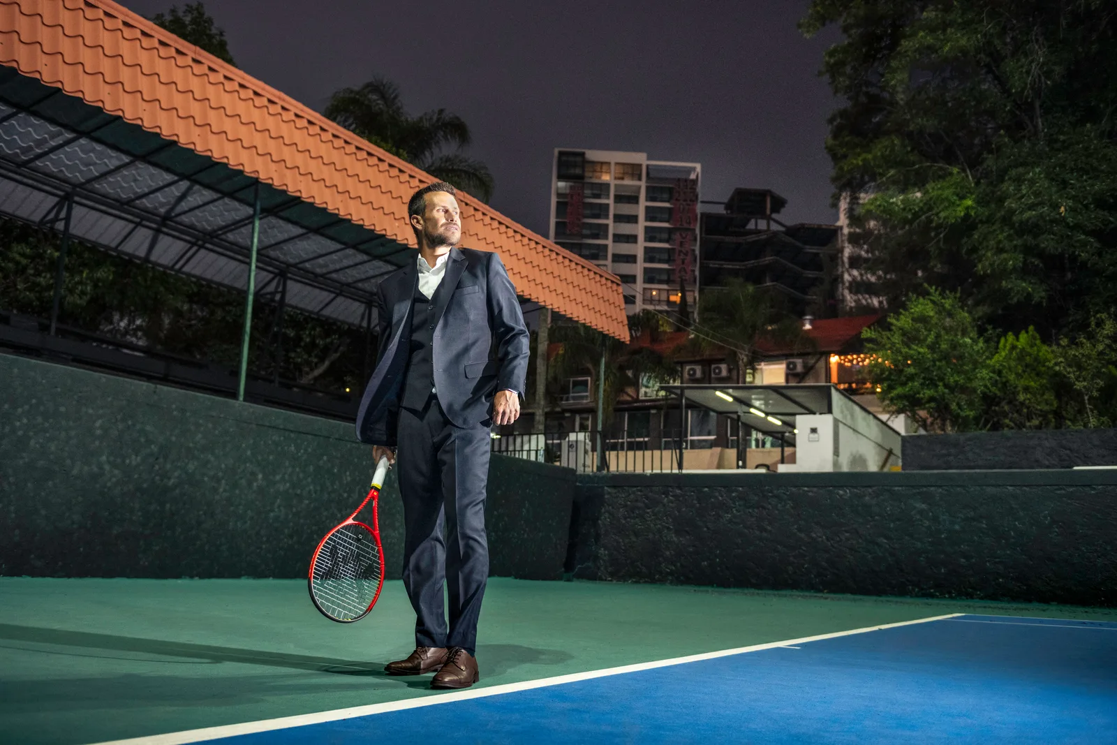 Miguel, in a suit and holding a racket, on a tennis court at night.
