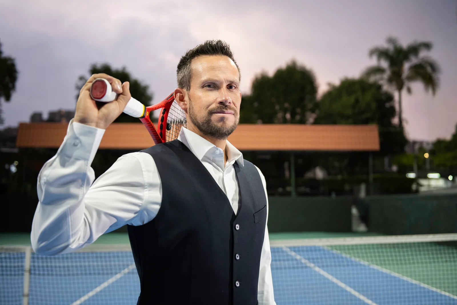 Miguel, in a waistcoat and white shirt, holds the racket on his shoulder on the court at dusk.